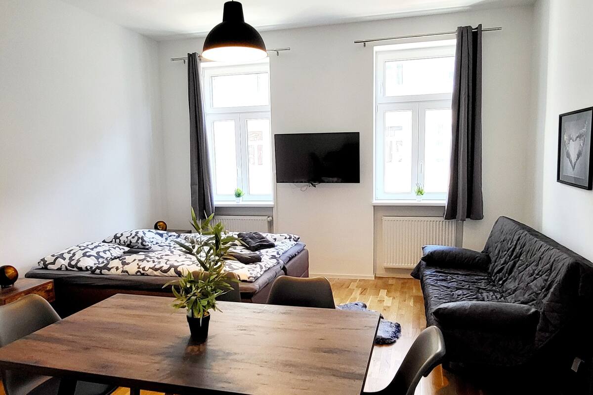 A modern living room with a wooden dining table, a sofa, and a TV mounted on the wall.