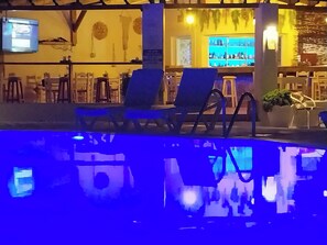 A pool area with lounge chairs and a bar area with a television screen.