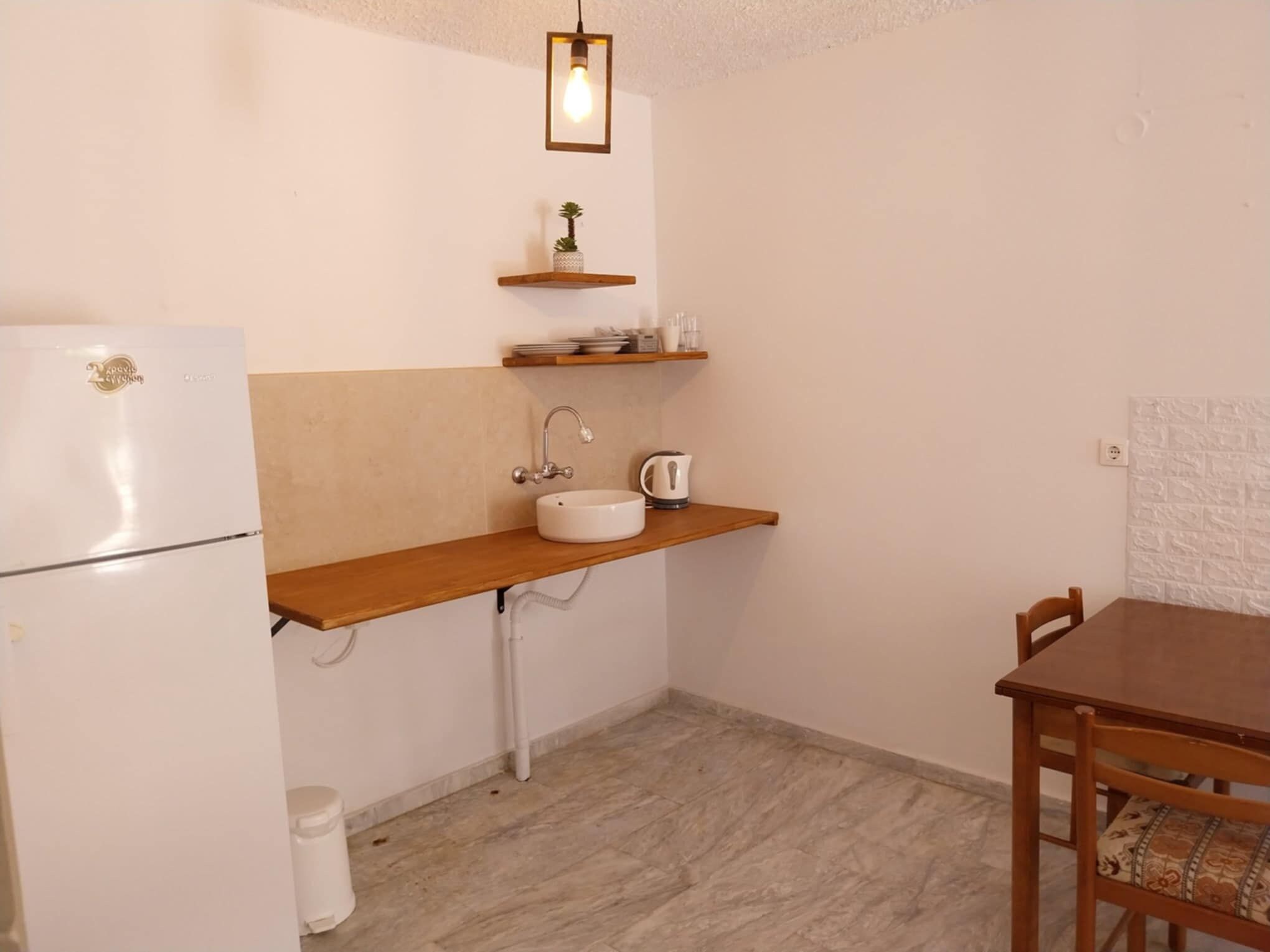 A compact kitchen with a white refrigerator, a sink, a wooden shelf with kitchen items, and a dining table with chairs.