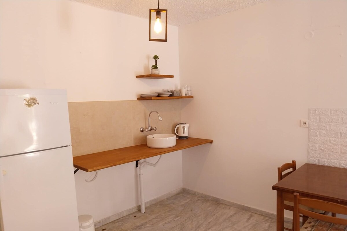 Una cocina compacta con un refrigerador blanco, un lavamanos, un estante de madera con utensilios de cocina y una mesa de comedor con sillas.