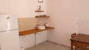 A compact kitchen with a white refrigerator, a sink, a wooden shelf with kitchen items, and a dining table with chairs.
