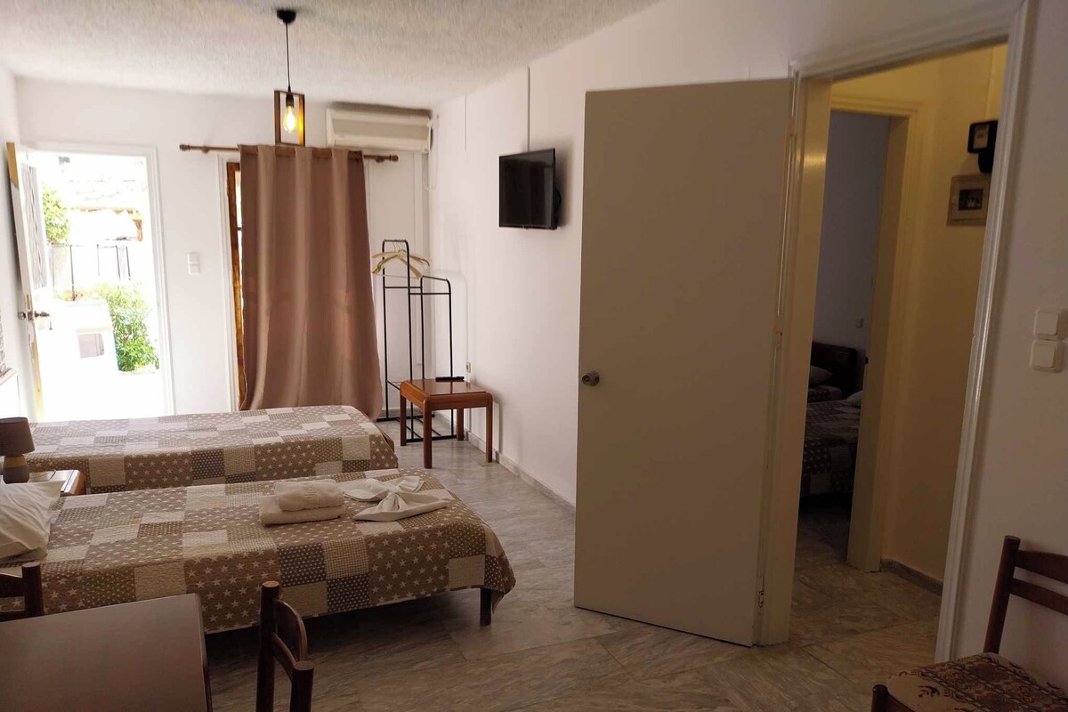 A room with two beds, a TV mounted on the wall, a door leading to another room, and a small table with a chair.