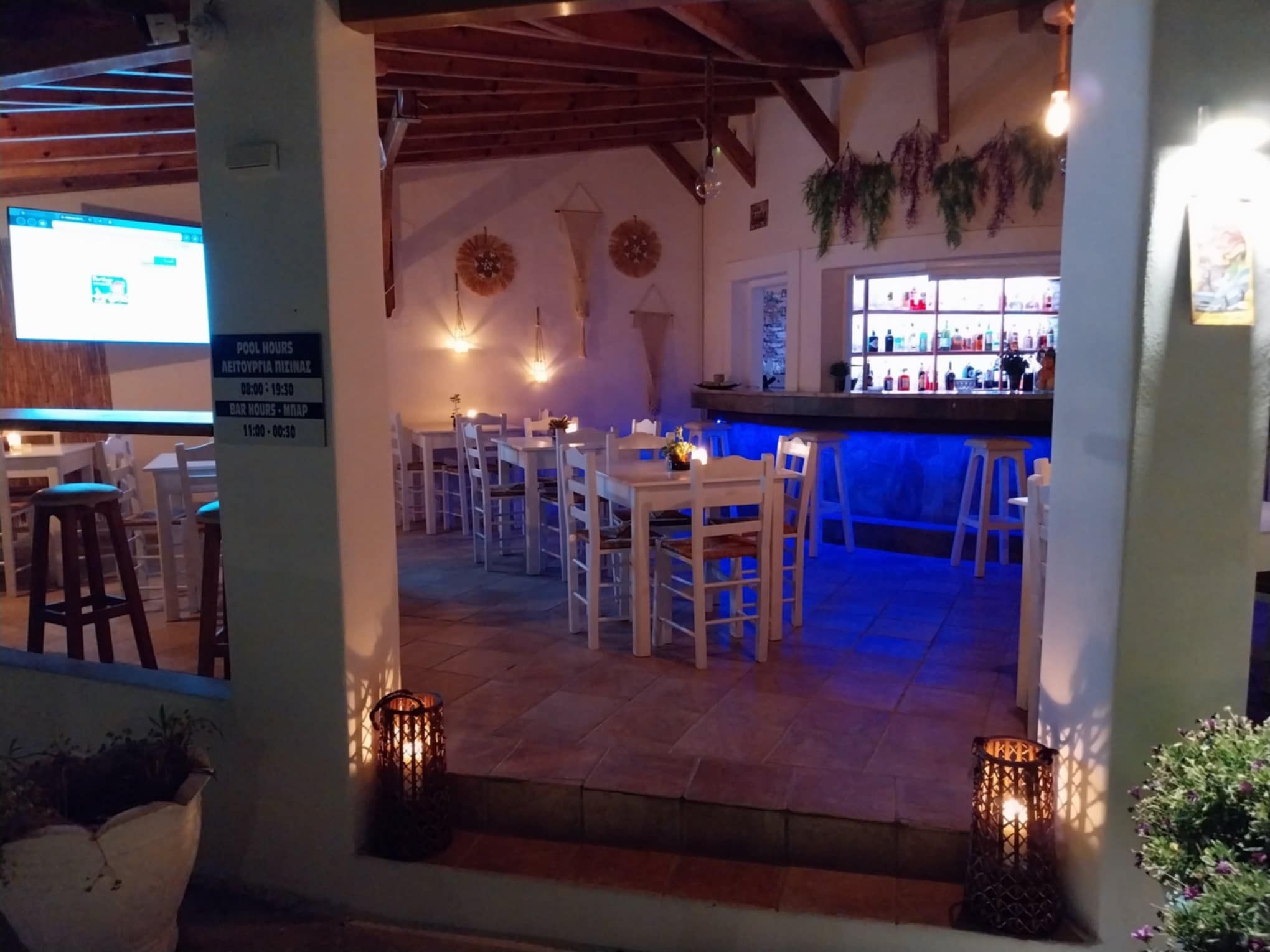 Un área de comedor al aire libre con mesas y sillas blancas, una barra con iluminación azul y adornos colgantes en la pared.