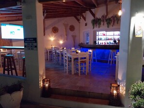 Un área de comedor al aire libre con mesas y sillas blancas, una barra con iluminación azul y adornos colgantes en la pared.