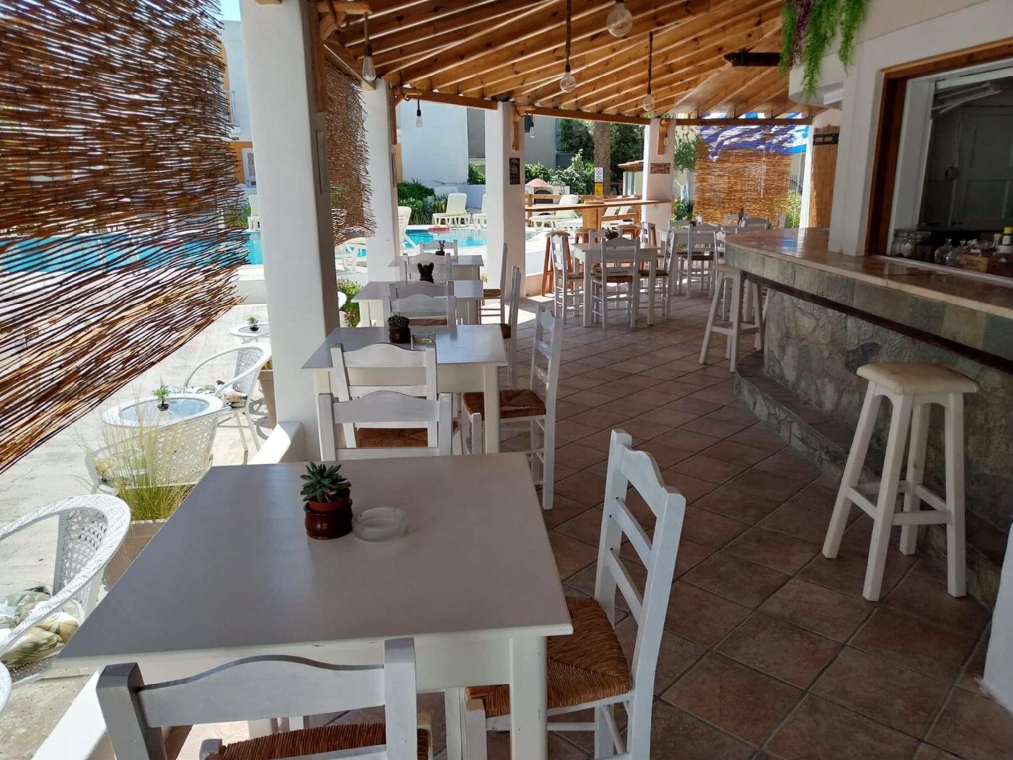A covered outdoor dining area with white tables and chairs, a stone counter, and a pool in the background.