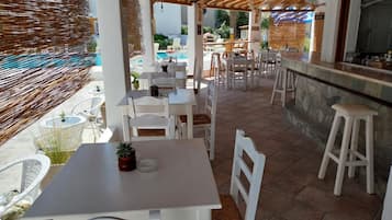 A covered outdoor dining area with white tables and chairs, a stone counter, and a pool in the background.