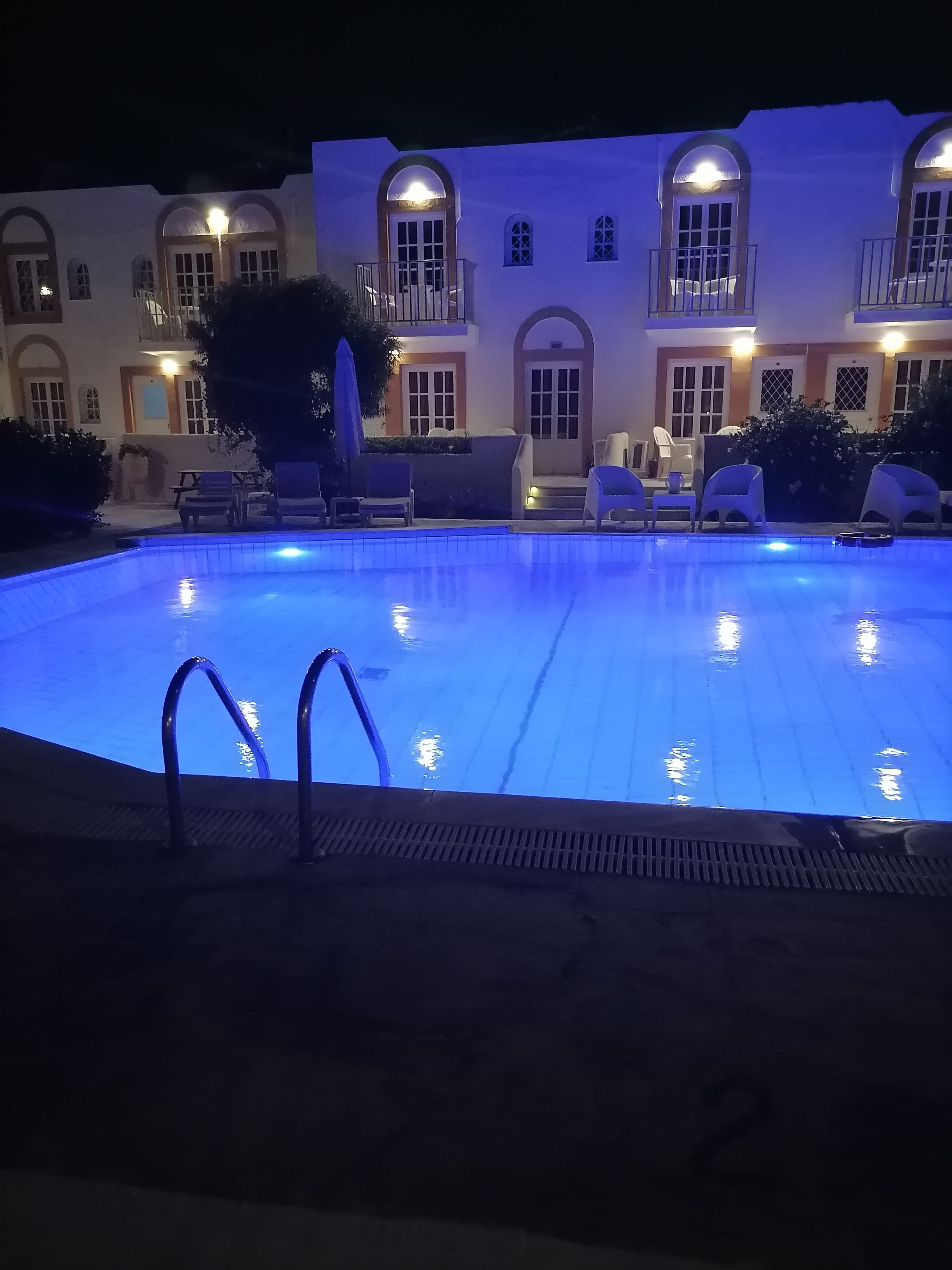 Piscina com superfície de água iluminada em azul, rodeada por espreguiçadeiras e um edifício com janelas e portas em arco.