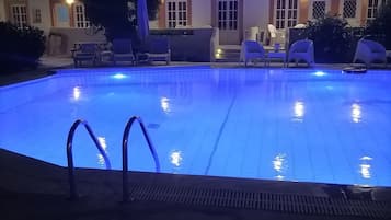 Piscina com superfície de água iluminada em azul, rodeada por espreguiçadeiras e um edifício com janelas e portas em arco.
