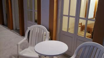 Un pasillo con sillas de plástico blancas y una pequeña mesa redonda, que conduce a una habitación con una cama y una mesita de noche.