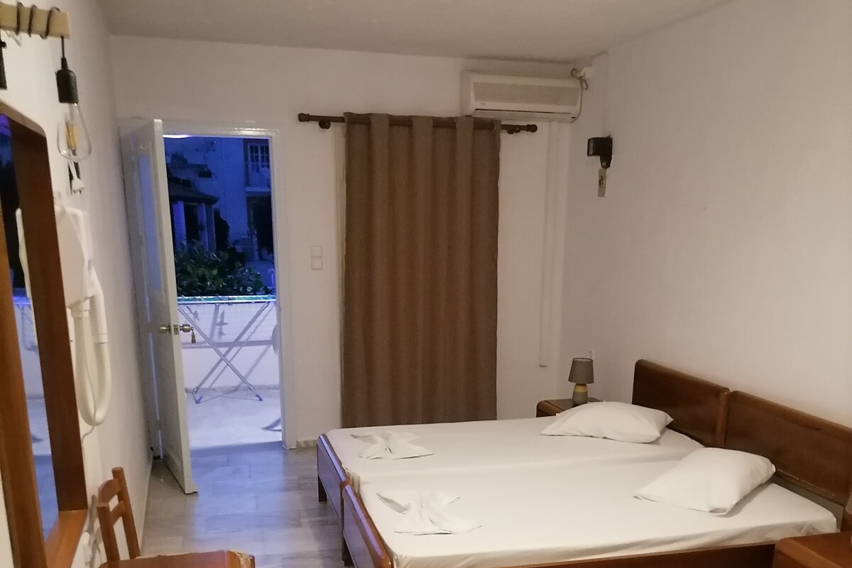A bedroom with a wooden bed, a desk, and a chair. There is a window with curtains, an air conditioning unit on the wall, and a view of the outside.