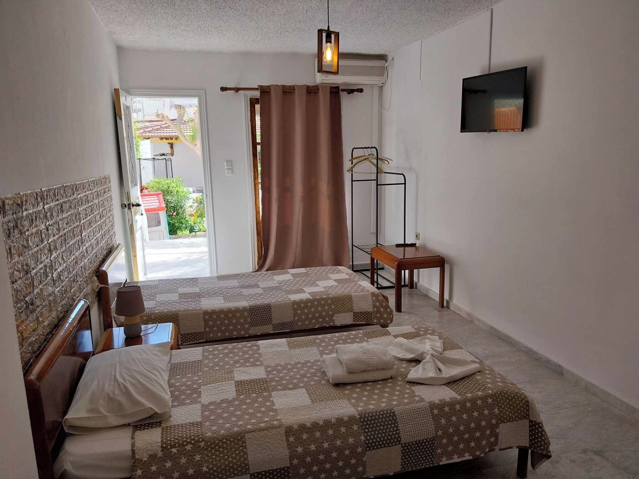 A small, clean room with two beds, a TV, a window with curtains, and a door leading outside.