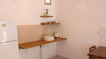 Eine kleine Küche mit einem weißen Kühlschrank, einer Holz Arbeitsplatte, einer Spüle und einem Wasserkocher. Oberhalb der Arbeitsplatte befinden sich zwei Holzregale, eines mit einer kleinen Pflanze, das andere mit ein paar Gegenständen.