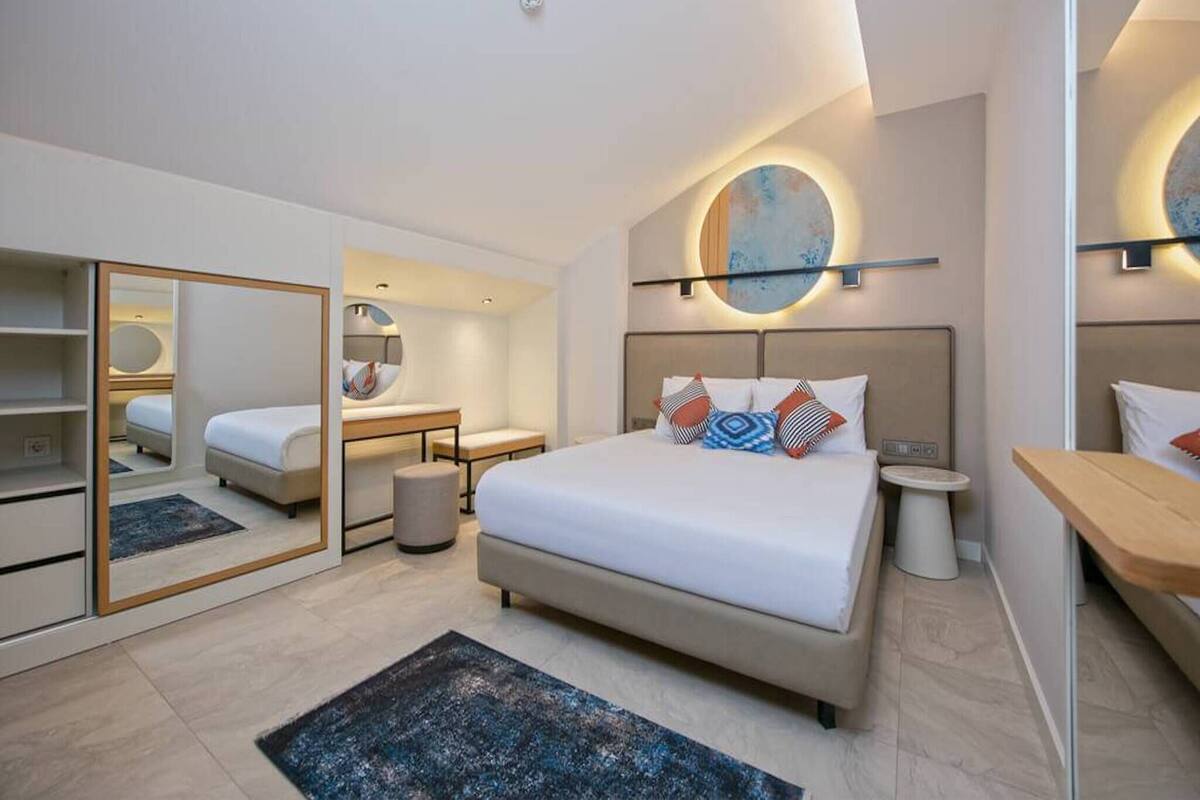 A modern hotel room with a large bed, bedside tables, a desk, a mirror, and a round wall light.