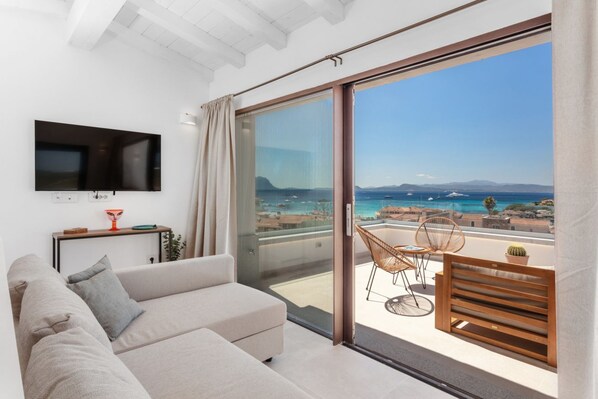A modern living room with a sofa, a TV mounted on the wall, and a balcony with a view of the sea.