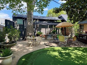 Eine Terrassenfläche mit Tisch und Stühlen, ein kleiner Garten mit Baum und Pflanzen, und ein Haus im Hintergrund.