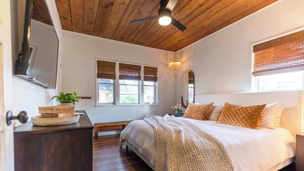 A bedroom with a wooden ceiling, a bed with white bedding, a bedside table, a mirror, and a window with blinds.