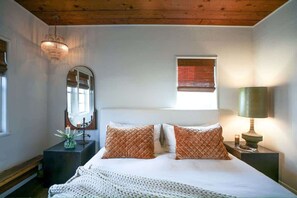 Ein Schlafzimmer mit Bett, Nachttisch, Lampe, Spiegel und einem Fenster mit Jalousien.