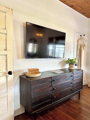 A room with a dark wooden dresser, a flat-screen TV mounted on the wall, a door with a light switch, and a potted plant on the dresser.