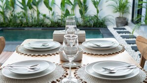 A dining table set with plates, cutlery, and glasses, surrounded by a pool and greenery. - Casa de Cavendish (Canggu)