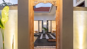 A modern hotel entrance with a wooden archway, a tiled roof visible above, and a well-lit interior with a bed and a small seating area. - Casa de Cavendish (Canggu)