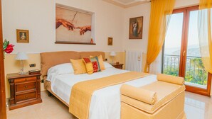 A bedroom with a large bed, a nightstand, a lamp, a painting, and a view of the outdoors.