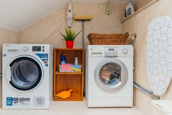 Two front-loading washing machines in a laundry room with a shelf holding cleaning supplies, a potted plant, and a laundry basket.