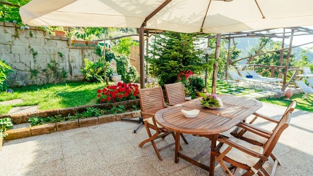 Une terrasse extérieure avec une table ronde en bois et des chaises, un grand parasol, et un jardin agrémenté de fleurs rouges et de verdure.