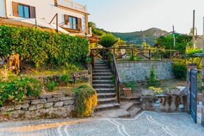 Una escala de pedra que condueix a un edifici amb un balcó, envoltat de vegetació i un camí de llambordes.