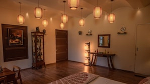 A well-lit room with wooden flooring, a bed with patterned bedding, a dining table with chairs, a cabinet with glass doors, and multiple hanging lamps. - Amã Stays & Trails Butterfly Villa, Deolali, Nashik (Nashik)