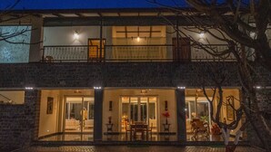 A two-story building with a balcony, a dining area with a table and chairs, and a large window. - Amã Stays & Trails Butterfly Villa, Deolali, Nashik (Nashik)