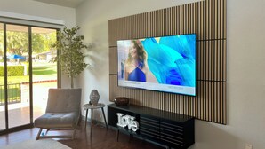 Uma sala de estar moderna com uma grande TV de tela plana presa a uma parede com lambris, uma poltrona cinza confortável, uma pequena mesa lateral com objetos decorativos e vista para uma área externa bem cuidada através de grandes portas de vidro.