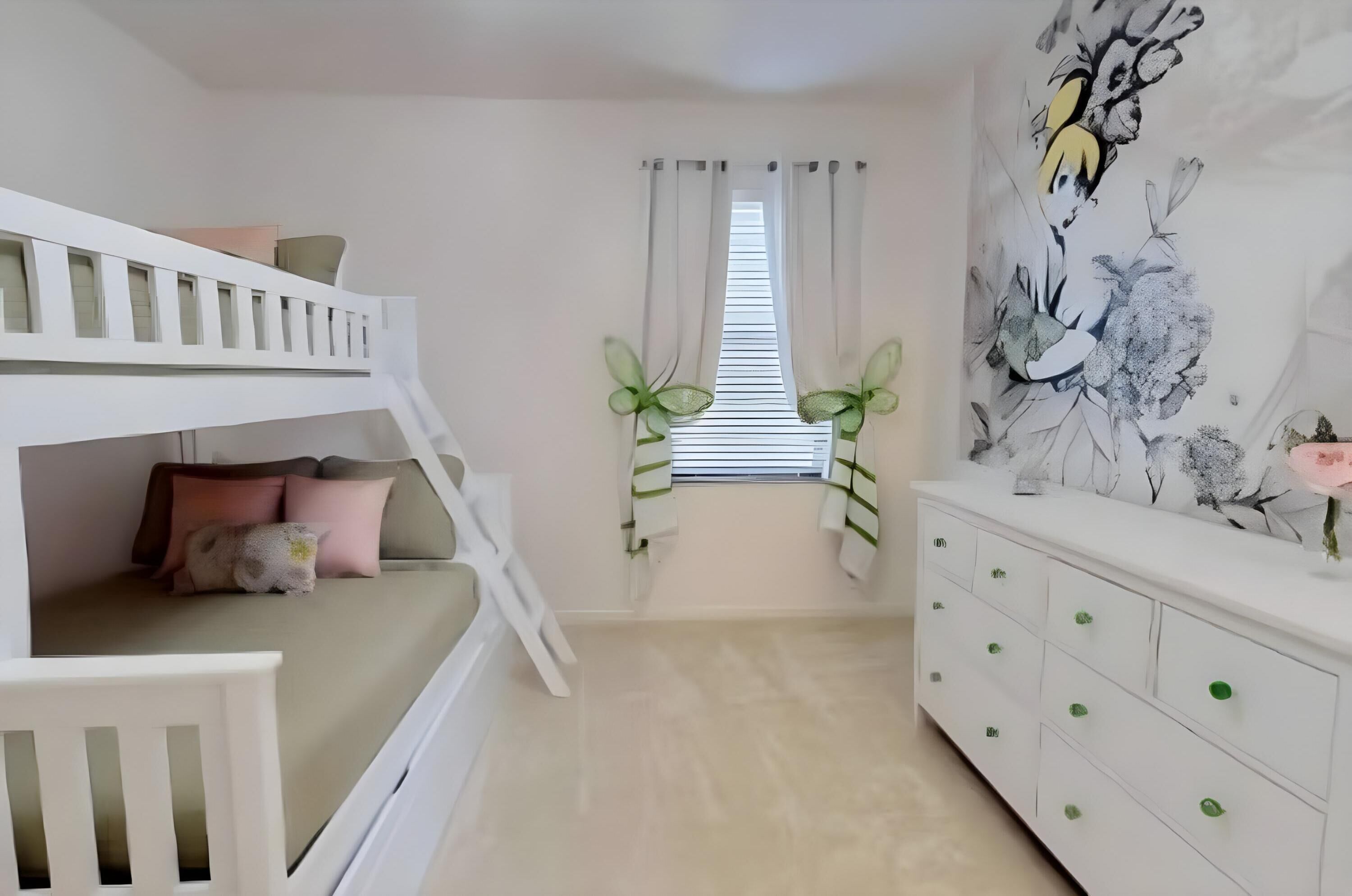 A child’s bunk bed with a pink pillow, a white dresser with green knobs, and a wall with a floral painting.