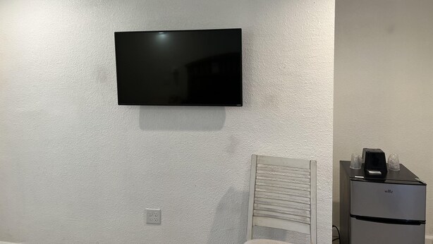 Quarto com televisão de tela plana montada, parede branca com textura, cadeira branca e um pequeno frigorífico.