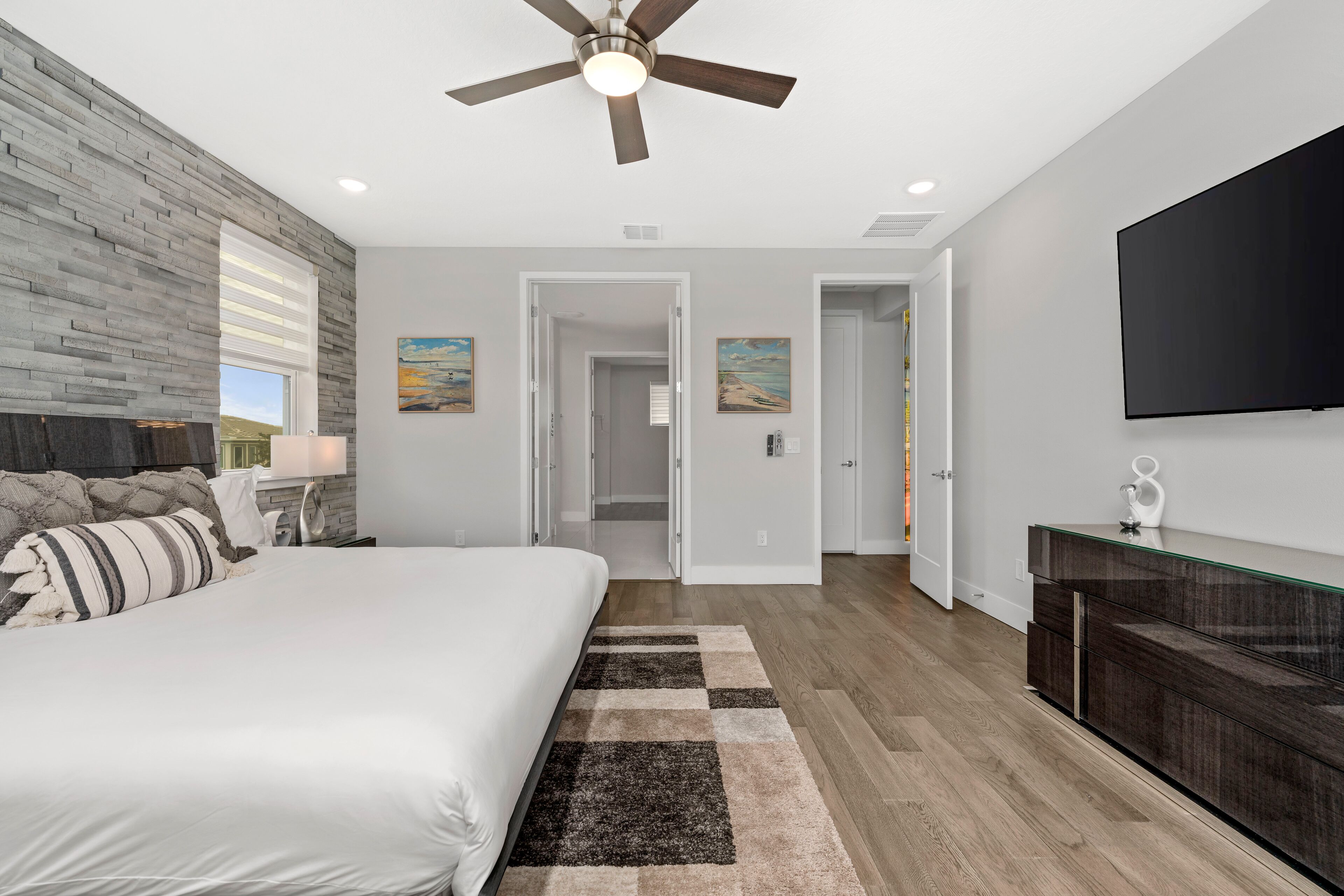 A modern bedroom with a large bed, a ceiling fan, a flat-screen TV, and a wooden floor.