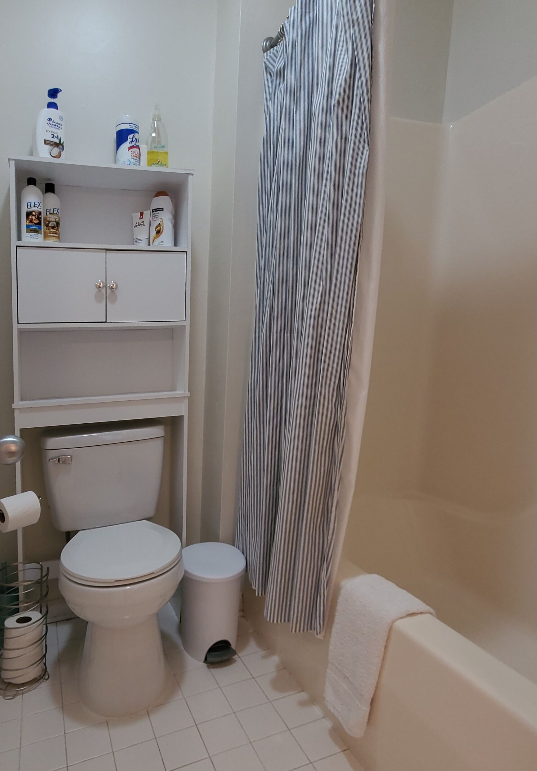 Un baño con inodoro, un cubo de basura, un estante con artículos de aseo y una cortina de ducha.