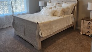 A neatly made bed with a wooden headboard, two bedside tables with lamps, and a window with sheer curtains. - Entire New home in Edmond, Oklahoma (Edmond)