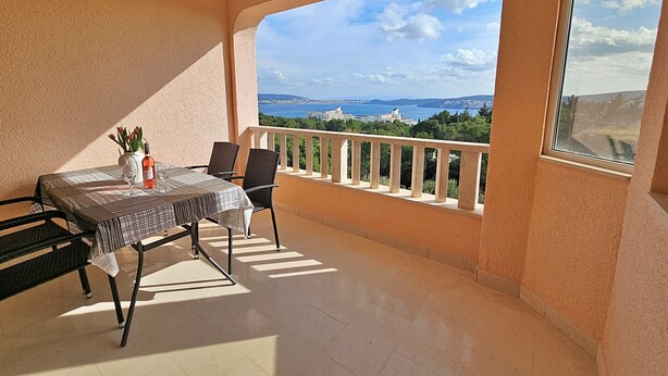 Un balcon avec une table dressée pour deux personnes, un vase rempli de fleurs et une bouteille de liqueur. Le balcon offre une vue panoramique sur la mer et les bâtiments éloignés.