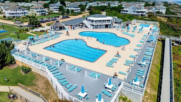 En stor swimmingpool med flere baner, omgivet af liggestole og parasoller, beliggende i et resortområde med huse i baggrunden.