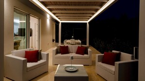 An outdoor patio with white sofas and red cushions, a table with a lamp, and a view of a dining area in the background. - Ecovilla Terra Di Luce 8 Emma Villas (Martina Franca)