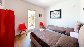 A room with two beds, a red chair, a red cabinet, and a window with a view of trees. - Ecovilla Terra Di Luce 8 Emma Villas (Martina Franca)