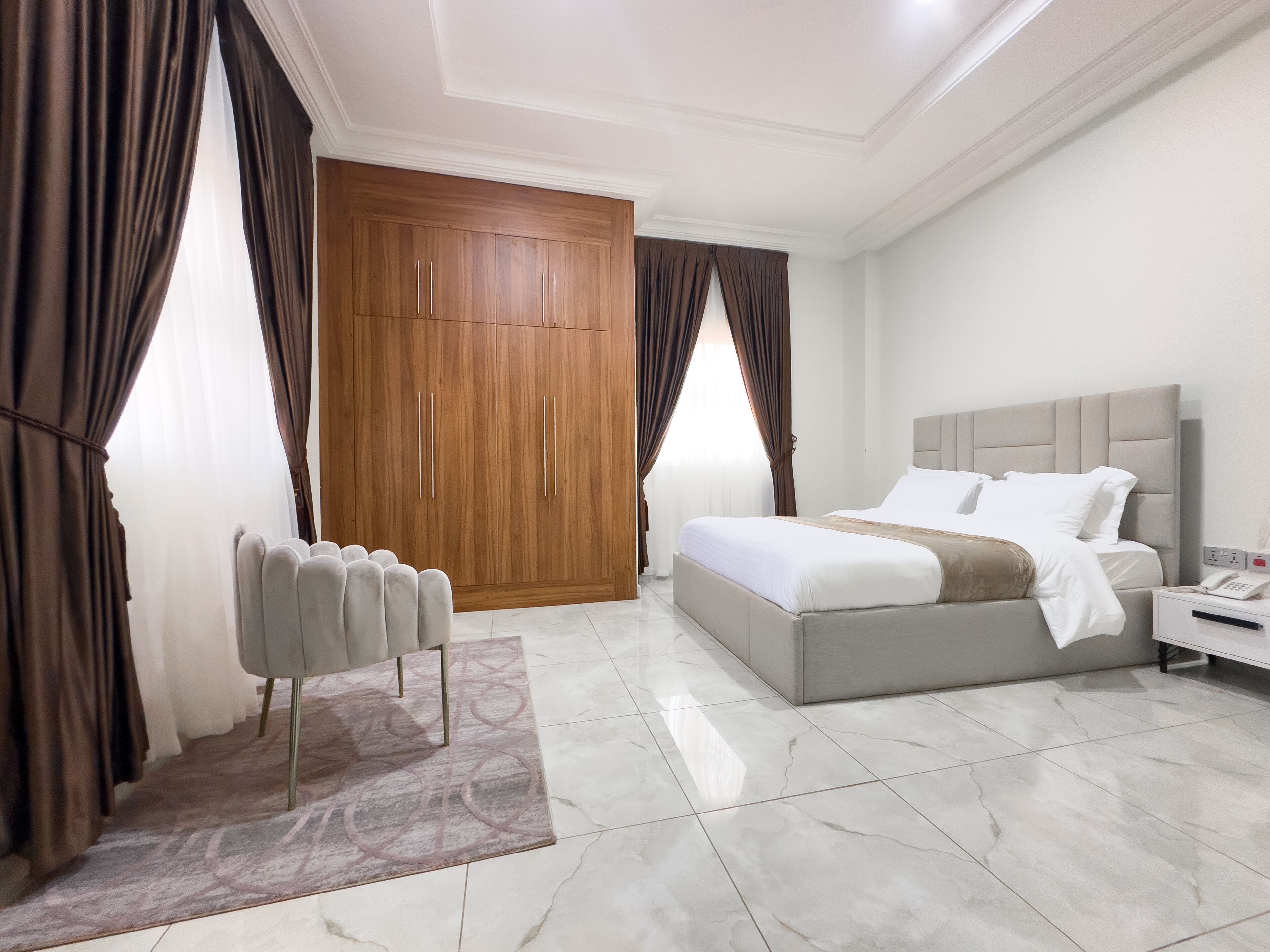 A modern hotel room with a large bed, a nightstand, a wardrobe, and a seating area with a chair and a small table.