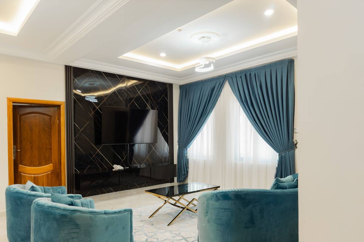 Een moderne woonkamer met een flatscreen televisie, twee blauwe fauteuils, een salontafel en een zwarte accentmuur met een geometrisch patroon.