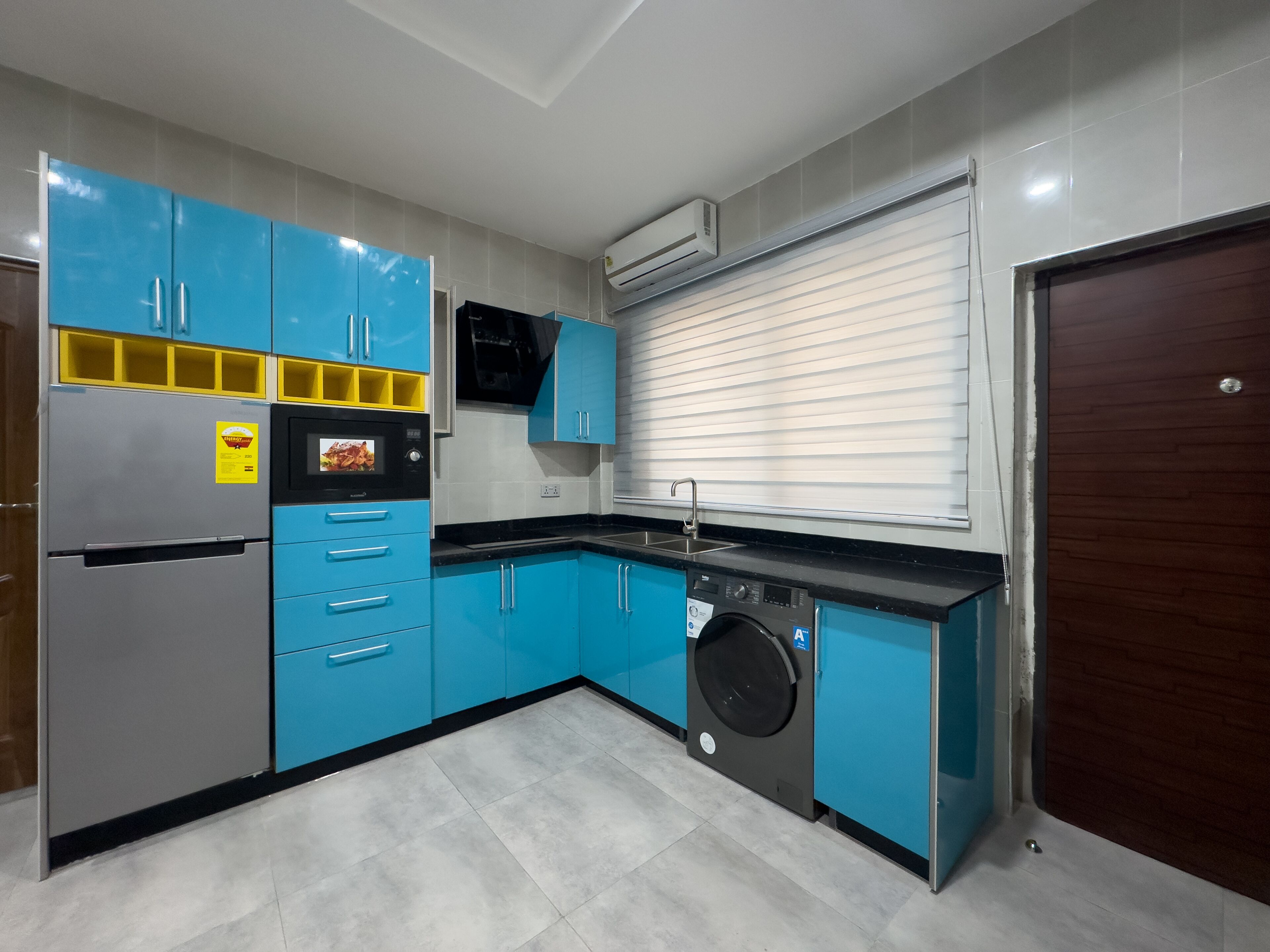 A modern kitchen with blue cabinets, a built-in microwave, a washing machine, and a large window with blinds.