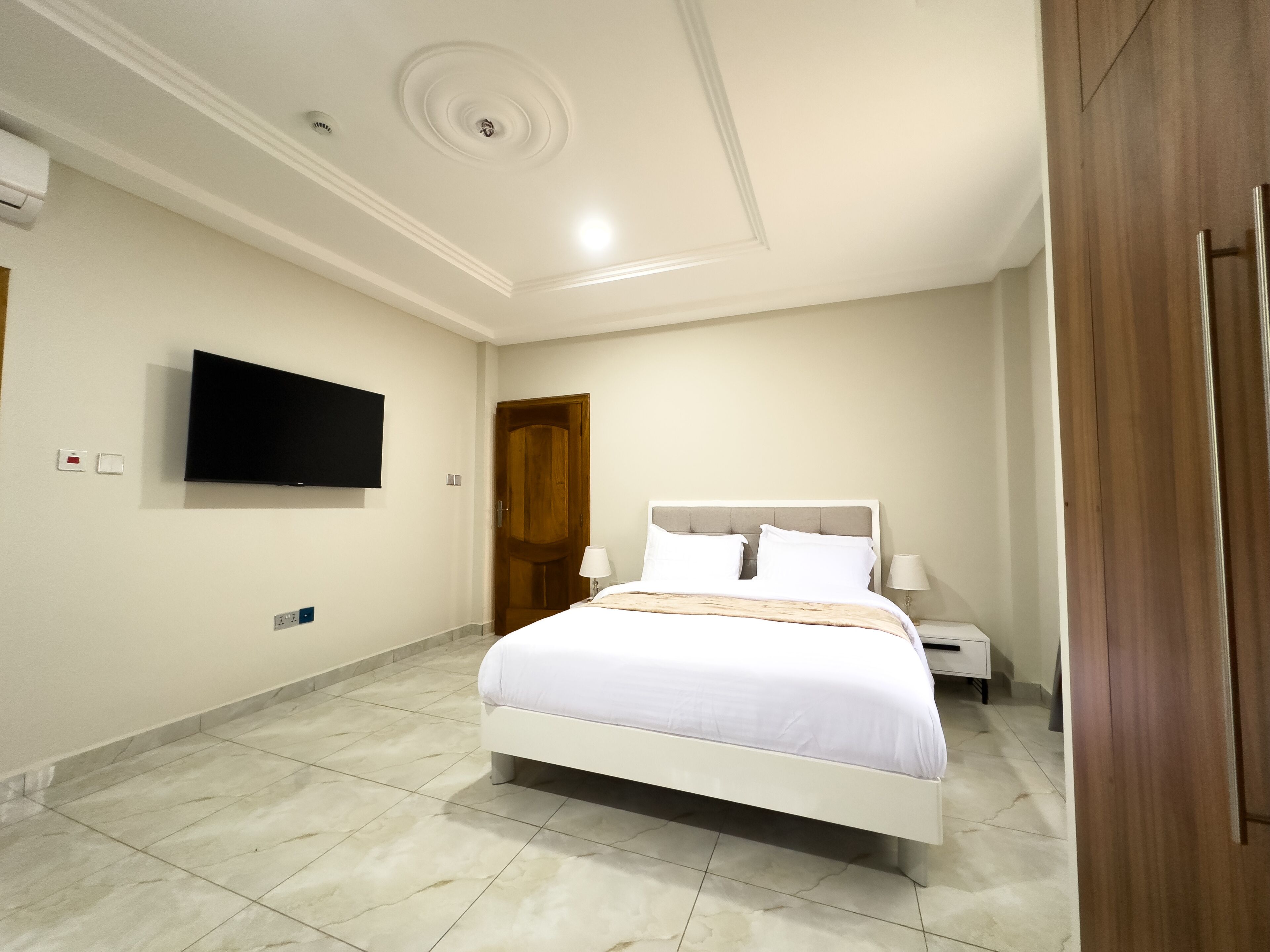 A bedroom with a bed, a TV mounted on the wall, a wooden door, and a nightstand with a lamp.