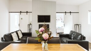 A modern living room with a wooden dining table, two black leather sofas, a flat-screen TV mounted on the wall, and a vase with flowers on the table. - Wallaby 2 Bed, 1 Bath Cottage - Private Parkland Setting with Lake View (North Maleny)