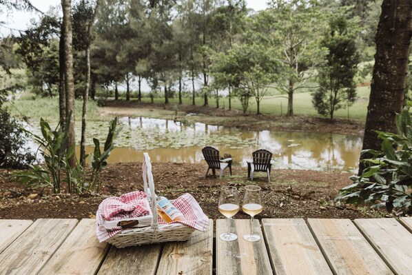 A wooden deck with a picnic setup including a basket, two glasses of white wine, and two chairs. - Wallaby 2 Bed, 1 Bath Cottage - Private Parkland Setting with Lake View (North Maleny)