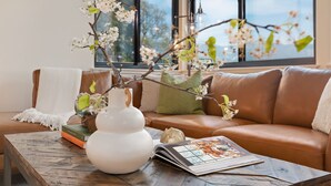 A cozy living room with a brown leather sofa, a white vase with branches, and a wooden coffee table with books and a magazine. - Panoramic Ocean and City Views at Luxury Home with Hot Tub (Santa Barbara)