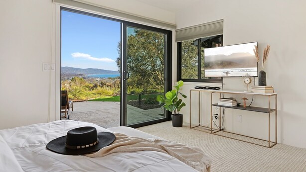 A bedroom with a large bed, a sliding glass door leading to a balcony, a TV, a desk with a plant, and a view of the outdoors. - Panoramic Ocean and City Views at Luxury Home with Hot Tub (Santa Barbara)