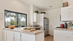 A modern kitchen with white cabinets, a stainless steel refrigerator, and a large window providing a view of trees. - Panoramic Ocean and City Views at Luxury Home with Hot Tub (Santa Barbara)