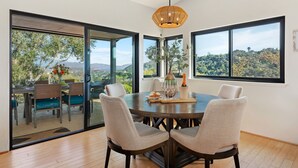 A dining area with a round table, four chairs, a chandelier, and large windows offering a view of trees and hills. - Panoramic Ocean and City Views at Luxury Home with Hot Tub (Santa Barbara)